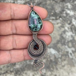 May include: A handcrafted pendant featuring a teardrop-shaped gemstone with green and pink hues, encased in copper wire. The wirework extends into a spiral design, with a curved tail. The pendant is approximately 2 inches long.