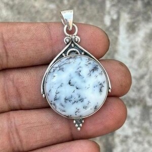 May include: A silver pendant with a large, round, white stone featuring black dendritic inclusions. The stone is set in a decorative silver frame with a bail for a chain. The pendant is held in a hand.