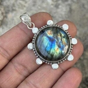 May include: A round pendant featuring a polished labradorite gemstone with iridescent blue and gold flashes. The stone is set in a silver-toned metal frame decorated with small circular accents around the perimeter and a textured bail at the top.