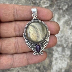 May include: A silver pendant featuring a large, round, iridescent labradorite stone at the center, accented by a smaller, teardrop-shaped purple gemstone at the base. The metal setting includes intricate wire-work detailing and a top bail loop.