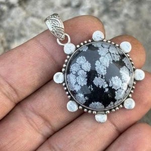 May include: A round pendant featuring a black and white snowflake obsidian stone set in a silver-toned metal frame. The circular stone displays natural white floral-like patterns against a dark background, surrounded by six small silver spheres.