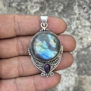 May include: A silver pendant featuring a large, round, iridescent labradorite gemstone with blue and gold flashes, accented by a smaller, teardrop-shaped purple amethyst stone at the base, all set within a decorative, ornate metal frame.