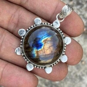 May include: A round pendant featuring a polished labradorite gemstone with iridescent blue and gold flashes. The stone is set in a silver-toned metal frame with small circular accents around the perimeter and a textured, ornate bail at the top.