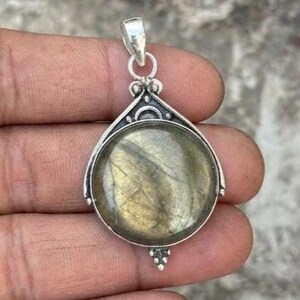 May include: Silver pendant with a large, round, iridescent gemstone. The stone has a mix of green, gold, and gray tones. The pendant has a decorative silver setting with a loop for a chain. The pendant is held in a hand.