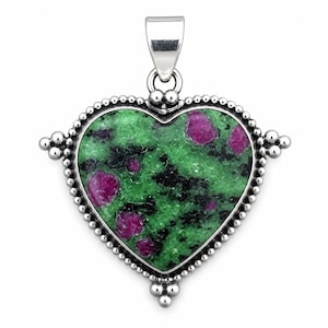 May include: A heart-shaped pendant featuring a green zoisite stone with embedded magenta ruby inclusions and black mineral patches. The stone is set in a polished silver frame with a beaded edge and a silver bail at the top for a necklace chain.