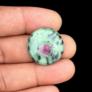 Può includere: Una gemma rotonda con una base verde chiaro, macchiata di nero e con un'inclusione centrale rosso intenso. La pietra è tenuta in mano su sfondo nero, mostrando la sua superficie lucida e i suoi motivi naturali.