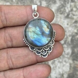 May include: A round labradorite pendant set in a silver frame. The gemstone displays iridescent blue and gold flashes across its surface. The silver setting features a decorative scalloped edge along the bottom half and a simple bail at the top.