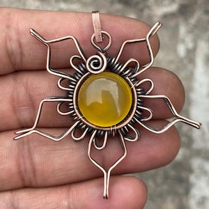 May include: A handcrafted copper pendant featuring a central, round, yellow gemstone. The gemstone is surrounded by a sunburst design made of copper wire. The pendant has a loop for a chain.