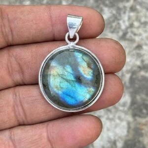 May include: A round pendant featuring a polished labradorite gemstone with iridescent blue and gold flashes, set in a smooth silver-toned metal frame with a simple triangular bail at the top for attaching to a chain or cord necklace.