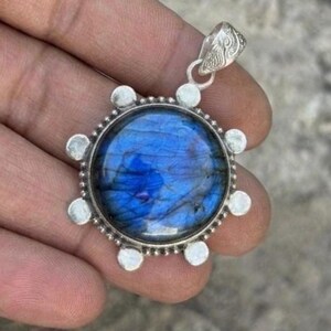 May include: A round pendant featuring a polished, iridescent blue labradorite gemstone set in a silver-toned metal frame. The circular setting is accented with small, smooth silver spheres around the perimeter and includes a decorative bail.