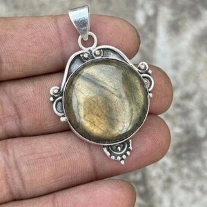 May include: A round labradorite gemstone pendant set in a silver-toned metal frame. The stone displays a muted golden-brown iridescence. The metal setting features decorative bead-like accents along the edges and a simple bail at the top.