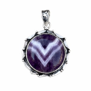 May include: A round pendant featuring a polished purple and white chevron amethyst stone set in a decorative sterling silver frame with a textured bail. The stone displays distinct V-shaped banding patterns in deep violet and opaque white tones.