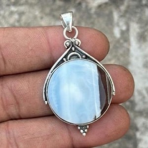May include: A silver pendant with a teardrop-shaped frame. The pendant features a large, round, light blue stone with a brown stripe. The top of the pendant has a loop for a chain, and the bottom has a decorative design.