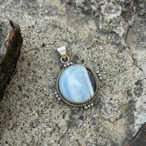 May include: A round silver pendant with a light blue and brown stone. The pendant has a twisted rope design around the stone and small silver beads around the edge. The bail is silver.