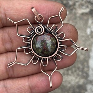 May include: Copper wire pendant with a central, round, green and red stone. The stone is encircled by a spiral of wire and surrounded by a sunburst design. The pendant has a small loop for a chain.