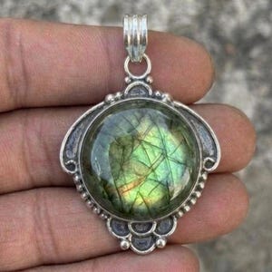May include: A round labradorite gemstone pendant set in a silver-toned metal frame. The stone displays a green and yellow iridescent sheen with dark internal inclusions. The metal setting features a decorative scalloped edge and a top bail.