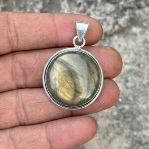 May include: A round pendant featuring a polished, iridescent labradorite gemstone set in a smooth silver-toned metal frame. The stone displays a muted greenish-grey hue with subtle golden undertones and a visible internal crystalline structure.