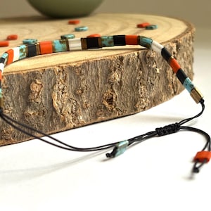 Peut inclure: Bracelet réglable avec un design linéaire. Il présente des perles rectangulaires orange, turquoise, noires et blanches. Le bracelet est monté sur un cordon noir avec une fermeture à nœud coulissant. Les perles mesurent environ 0,6 cm de long.