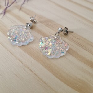 Peut inclure: Une paire de boucles d'oreilles argentées irisées en forme de coquillages avec des paillettes incrustées dans la résine. Les boucles d'oreilles ont une fermeture à tige.
