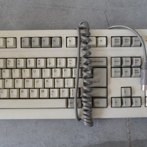 May include: A white computer keyboard with a coiled gray cable attached. The keyboard has a label that reads "MITLASER".