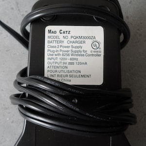 May include: Black power adapter with a white label that reads "Mad Catz Model No. PQKM3000ZA Battery Charger Class 2 Power Supply Plug-in Power Supply for Use with 8256 Wireless Controller Input: 120V~60Hz Output: 9V 120mA Attention Pour Utilisation L'Int Rieur Seulement In China" and a UL safety certification mark.