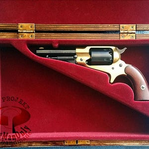 Pode incluir: Um revólver dourado com cabo de madeira repousa numa caixa de madeira forrada a veludo vermelho. A caixa tem uma dobradiça de latão e está aberta.