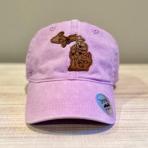 May include: A lavender baseball cap with a brown leather patch in the shape of the state of Michigan. The patch features a floral design. The cap has a curved brim and a small Detroit sticker.