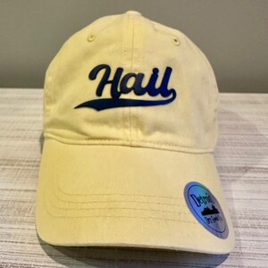 May include: A light yellow baseball cap with the word "Hall" in dark blue script across the front. The cap has a curved brim and a small sticker on the side. The cap is made of a soft material.
