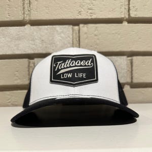May include: A white and black trucker hat with a black brim. The hat features a black and white patch with the words "Tattooed LOW LIFE" in a stylized font.