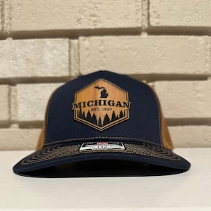 May include: A navy blue and tan trucker hat with a wooden patch featuring the state of Michigan and the text "MICHIGAN EST. 1837". The hat has a curved brim and a mesh back.