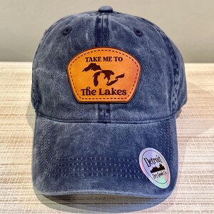 May include: A blue denim baseball cap with a leather patch that reads "TAKE ME TO The Lakes". The patch features a silhouette of the Great Lakes. A small sticker with the text "Detroit Dry Goods Co." is on the brim.