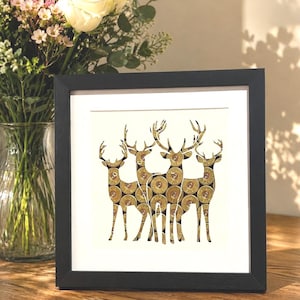 May include: A framed art print depicting five deer. The deer are formed from gold-coloured bullet casings, set against a white backdrop. The black frame provides a contrasting border. A vase of flowers is in the background.
