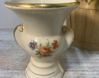 May include: A small, white ceramic vase with a gold rim and floral design. The vase has two handles and a crackled glaze.