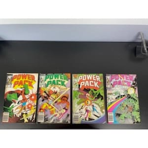 May include: Four vintage Power Pack comic books. The covers feature vibrant illustrations with the title "Power Pack" in bold lettering. The comic book covers have a variety of colors, including red, green, yellow, and purple. The comics are displayed on a dark surface.