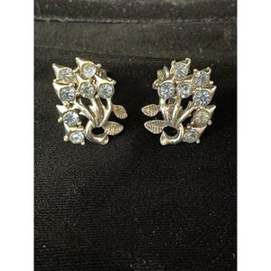 May include: A pair of silver-toned clip-on earrings. Each earring features a floral design with clusters of light blue rhinestones. The earrings have a detailed, vintage aesthetic, with a classic clip-on mechanism.