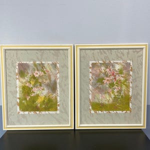 May include: Two framed floral watercolor paintings. Each features a central composition of pink and yellow flowers with green foliage against a textured background. The frames are white with a yellow inner edge.