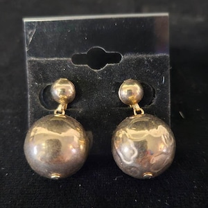 May include: A pair of silver-toned ball drop earrings. Each earring features a small, round stud connected to a larger, textured sphere. The earrings are displayed on a black card.