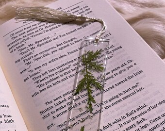 May include: A clear bookmark with a pressed green fern inside. The bookmark is rectangular and has a silver tassel attached to the top. The bookmark is resting on a page of a book with text that reads "The next patient Hutton looked at her..."