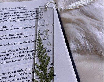 May include: A clear bookmark with a pressed green plant and white flowers inside. The bookmark has a white tassel attached to the top. The bookmark is laying on top of an open book with text visible. The text reads "THING LASTS FOREVER" and "my patients that way, Kat vowed."