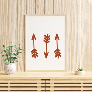 May include: A framed print with three stylized arrows in a terracotta color on a white background. The arrows are pointing in different directions. The print is hanging on a wood wall.
