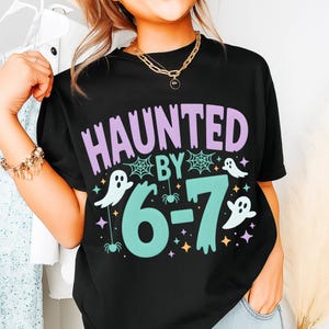 May include: Black t-shirt featuring the text "HAUNTED BY 6-7" in purple and teal, with ghost and spider illustrations. A fun Halloween or birthday-themed design. The t-shirt is a casual, comfortable style.