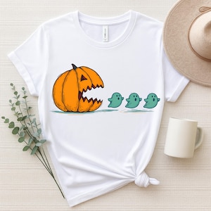 May include: White t-shirt with a Halloween graphic. An orange pumpkin with a jagged mouth is chasing three teal ghosts. The tee shirt has a crew neck design and is made of a soft material.