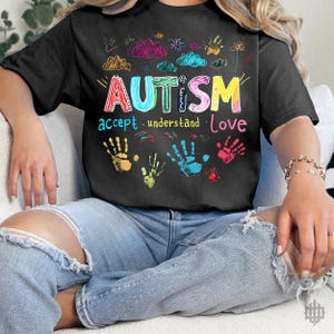 Puede incluir: Camiseta negra con un gráfico colorido que dice "AUTISMO aceptar. entender. amar" y presenta huellas de manos y nubes.