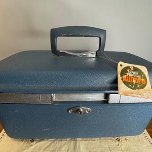 May include: A blue hard-shell train case with a textured finish, a top handle, and a silver-toned metal band around the center. A circular tag attached to the handle features the text Royal Traveller Aurora in orange and white lettering.