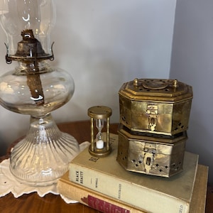 May include: An antique oil lamp with a clear glass globe and brass accents sits next to a small brass hourglass and two brass boxes. The items are placed on top of two vintage books with gold lettering.