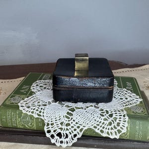 May include: A small, square, black leather box with a smooth, dark finish and a centered, metallic gold-toned cube knob on the lid. The box features visible stitching along the edges and a structured, compact design with a flat top and base.