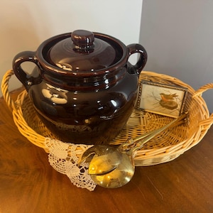 May include: A dark brown ceramic pot with a lid and handles sits in a woven basket. A small framed picture of a bird and a gold-colored ladle are also in the basket. The pot is a classic kitchenware item.