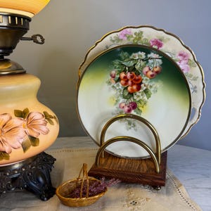 May include: Two decorative plates with floral and cherry designs, displayed in a wooden and brass plate holder. A small woven basket with purple flowers sits nearby. A vintage lamp with a floral design is in the background.