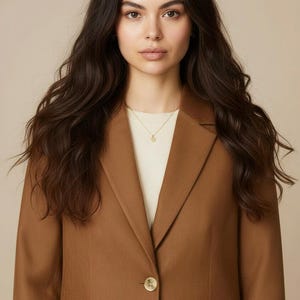 May include: A woman wearing a brown blazer over a white top. The blazer has a single button closure and a notched lapel. A gold necklace is visible. The woman has long, dark hair.