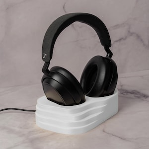 Könnte beinhalten: Schwarzer Over-Ear-Kopfhörer mit grauem Bügel auf einem weißen, mehrstufigen Ständer. Die Kopfhörer haben große, runde Ohrmuscheln und ein schlankes Design. Ein schwarzes Kabel geht vom Ständer ab.